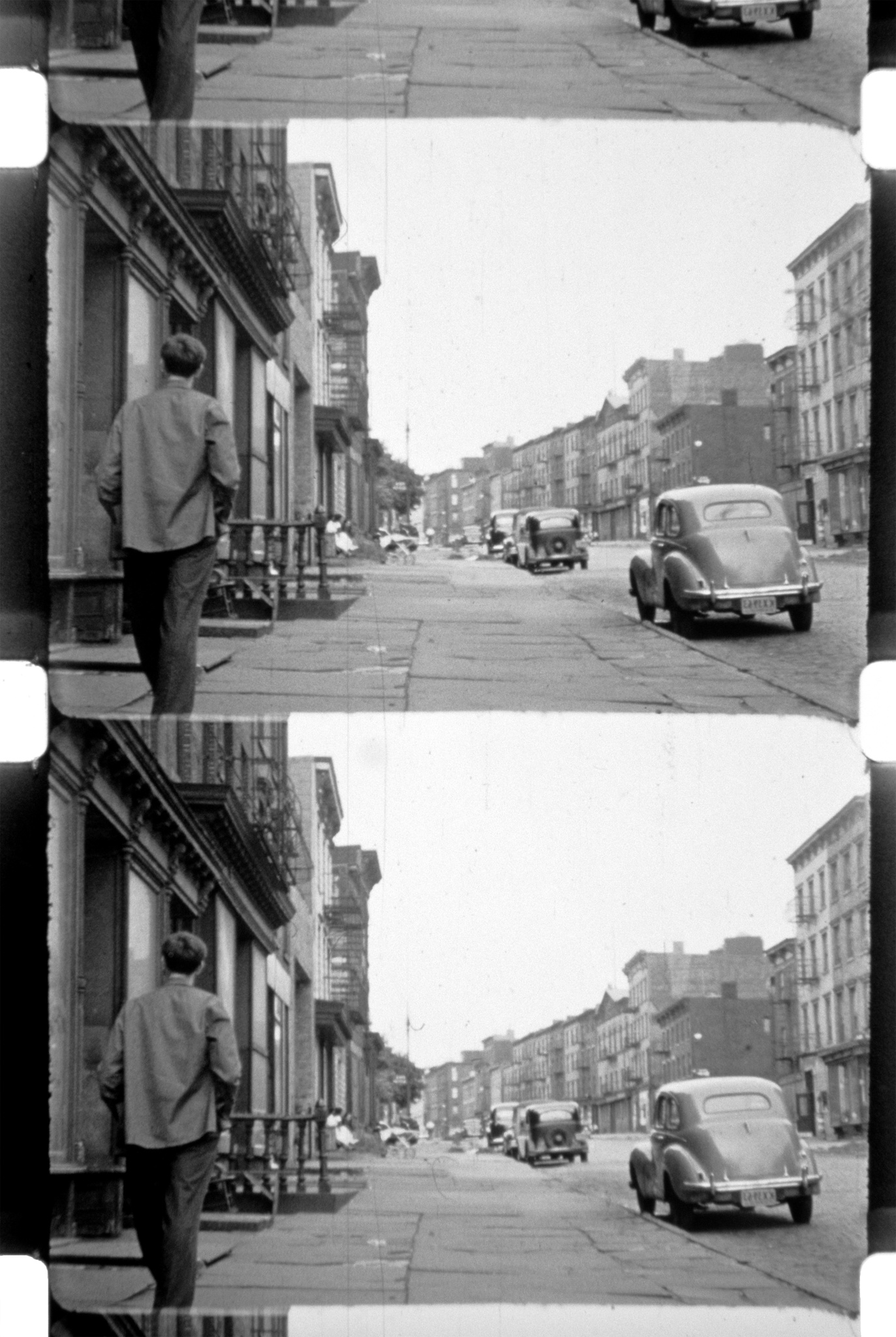 A black and white stereoscopic photograph showing a man standing on the sidewalk of a city street lined with buildings and parked cars from an earlier era, with more cars visible in the distance.