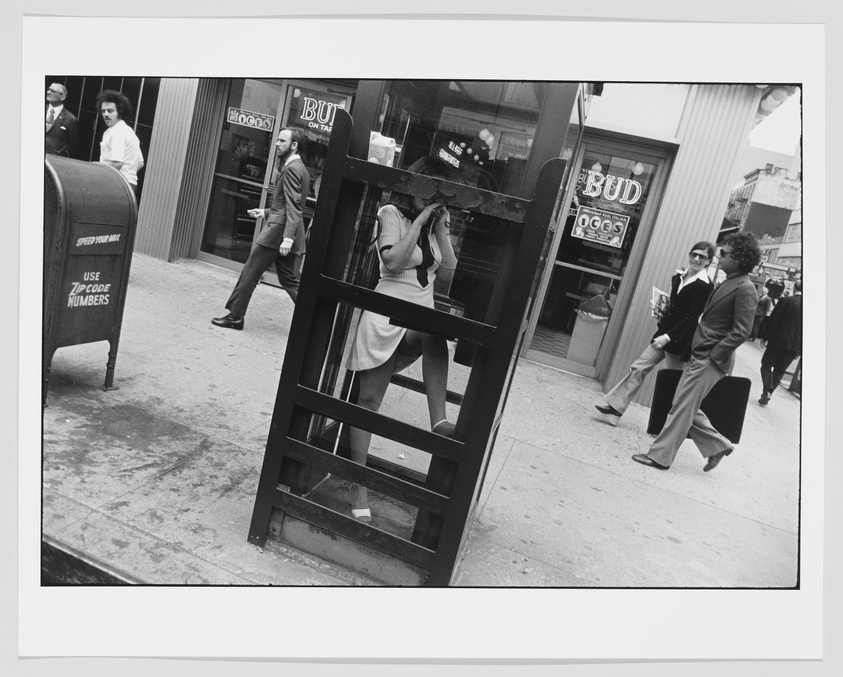 A black and white street photograph capturing a moment in time with pedestrians walking by. In the foreground, a person is partially obscured by a public telephone booth, with only their lower half visible. A mailbox with the text "USE ZIP CODE NUMBERS" is on the left, and a bar with a neon "BUD" sign is in the background. The scene suggests a bustling urban environment, possibly from a past decade given the fashion and hairstyles of the individuals in the frame.